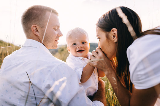 Walk beautiful young family in white clothes with a young son blond in mountainous areas with tall grass at sunset. Parents from both sides embracing son, hugging. family - this is happiness