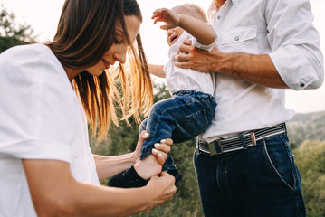 Obraz premium Walk beautiful young family in white clothes with a young son blond in mountainous areas with tall grass at sunset. Mother keeps son in her arms, hugging. family - this is happiness