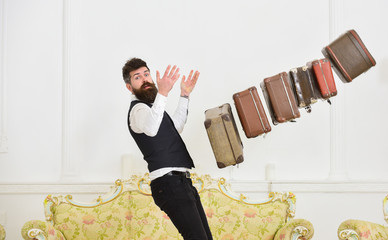Baggage insurance concept. Porter, butler careless, dropping pile of vintage suitcases. Man with beard and mustache in classic suit delivers luggage, luxury white interior background.