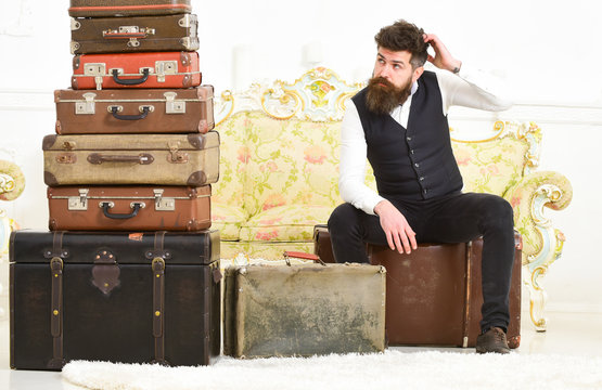 Luggage and relocation concept. Macho elegant on tired face sits, exhausted at end of packing, near pile of vintage suitcases. Man with beard and mustache packed luggage, white interior background.