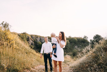 Walk of beautiful young family in white clothes with a young son blond in mountainous areas with tall grass at sunset. Dads carries his son on his shoulders. family - this is happiness