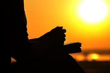 silhouette of the hands of woman praying to God in the nature witth the Bible at sunset, the...