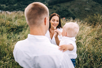 Walk beautiful young family in white clothes with a young son blond in mountainous areas with tall grass at sunset. Mother keeps son in his arms, hugging. family - this is happiness