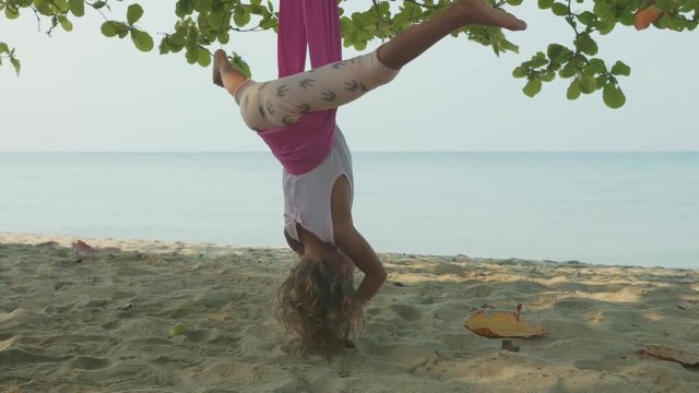 Little cute child girl doing yoga exersice with hammock in the beach.