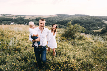 Naklejka na ściany i meble Walk beautiful young family in white clothes with a young son blond in mountainous areas with tall grass at sunset. Mother keeps son in his arms, hugging. family - this is happiness