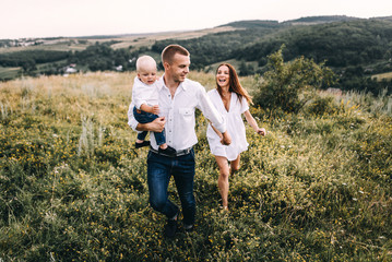 Fototapeta na wymiar Walk beautiful young family in white clothes with a young son blond in mountainous areas with tall grass at sunset. Mother keeps son in his arms, hugging. family - this is happiness