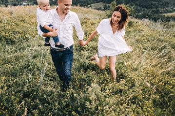 Walk beautiful young family in white clothes with a young son blond in mountainous areas with tall grass at sunset. Mother keeps son in his arms, hugging. family - this is happiness