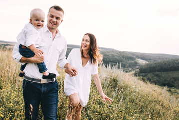 Walk beautiful young family in white clothes with a young son blond in mountainous areas with tall grass at sunset. Mother keeps son in his arms, hugging. family - this is happiness