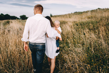 Walk beautiful young family in white clothes with a young son blond in mountainous areas with tall grass at sunset. Mother keeps son in his arms, hugging. family - this is happiness