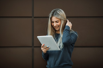 A young pretty girl is surfing on the internet on a tablet in the street on a brown modern background.