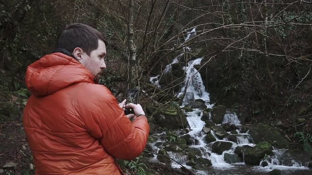 Shot from behind of tourist shooting video of a small waterfall in a mountains. Fantastic views of nature.