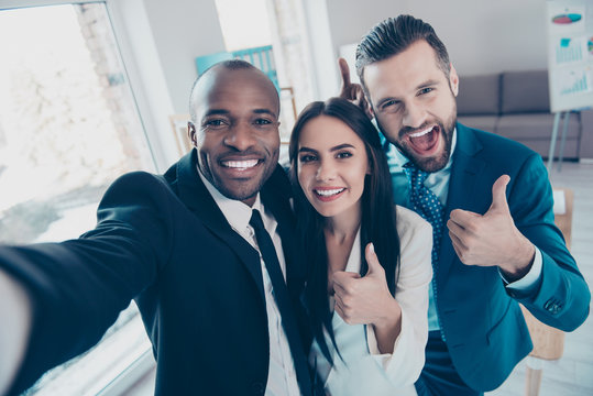 Three funny, cheerful friends having free time,  showing like symbol, afroamerican black man with bristle shooting selfie on front camera of smart phone, together standing in workplace, workstation