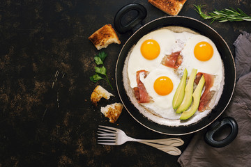 Breakfast with avocado, fried eggs, bacon, toast. Dark rustic background, top view, copy space.