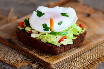 Delicious poached egg sandwich. Simple poached egg on rye bread slice with fresh vegetable mix and parsley. Healthy breakfast idea. Rustic wooden background. Closeup
