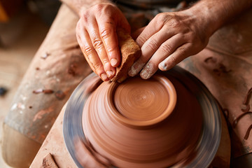 Adult male potter master modeling the clay plate on potter's wheel. Top view, closeup, hands only.