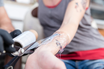 Close-up of the hands of a skilled tattoo artist wearing sterile black gloves while wrapping the forearm of a tattooed client
