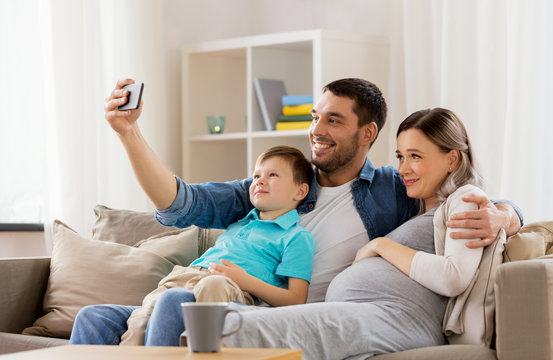 family, pregnancy and technology concept - happy pregnant mother, father and little son taking selfie with smartphone sitting on on sofa at home