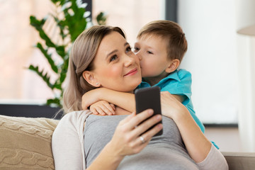 family, emotions and pregnancy concept - little son kissing happy pregnant mother with smartphone at home