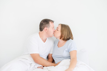 Kissing cute couple in sleepwear on the bed. Husband holding hand on stomach of his pregnant wife. Happy and loving family morning concept. Soft selective focus. Space for text.