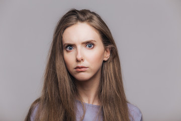 Headshot of beautiful female with blue eyes and pure skin, looks seriously and angrily at camera, has dark straight hair, isolated over grey background. Horizontal shot of lovely young woman indoor