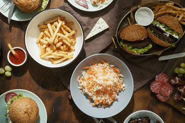 Dinner outdoor table with burger, french fries, salad and snacks on wooden table, top view