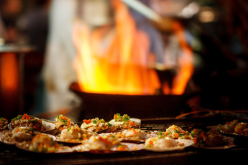 Grilled scallops sold at a street market stall with a pan covered with flames on the background....