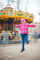 A photograph of a child's rest on nature in the spring. A child girl in a bright pink jacket is eating sweet cotton wool against the backdrop of rides, carousels. The girl jumps for joy