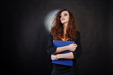 Brunette curly haired girl in black office jacket with skirt, on bra with folder of documentts at hands on studio against black background. Sexy businesswoman.