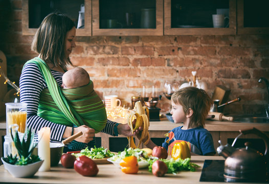 Happy Young Family, Beautiful Mother With Two Children, Adorable Preschool Boy And Baby In Sling Cooking Together In A Sunny Kitchen.