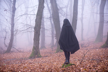Girl in black hood standing on a stump deep in a dark forest. Waiting for magic to happen. Thick fog all around. Scary autumn scene
