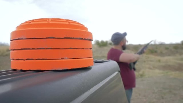 A young man shoots target practice at the gun range with orange clay pigeons in the foreground in slow motion. The overweight caucasian male uses a 12 gauge pump action shotgun to shoot skeet.
