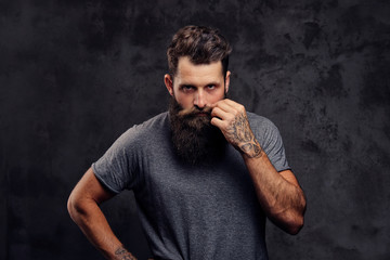 Portrait of a hipster with full beard and stylish haircut, dressed in a gray t-shirt, stands with a thinking look in a studio on a dark background.