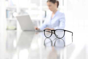 eyeglasses leaning on desk and woman working on computer at office in background