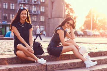 Two women tourists, relaxing after a walk in the sights, look in a map, sitting on stone steps against a background of bright sunset.