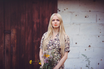 Fototapeta premium Beautiful young woman in a summer dress with wildflowers on a wall background with holes from bullets looking at the camera. aggression and military actions, occupied territories