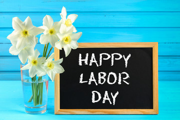 Chalkboard with the text: Happy Labor Day, May 1. White flowers of daffodils on a blue wooden table.