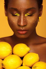 Cheerful young african woman with yellow makeup on her eyes. Female model against yellow background with yellow lemons.