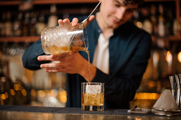 Brunet bartender pouring an alcoholic drink into a glass with ice cube