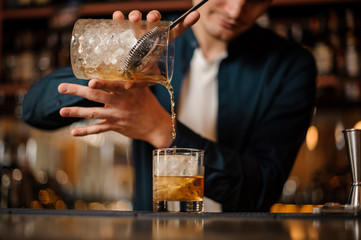 Brunet bartender pouring an alcoholic drink into a glass with a big ice cube