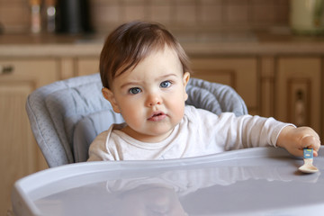 Hungry little baby sitting in the high chair at kitchen holding spoon in his hand, looking at camera
