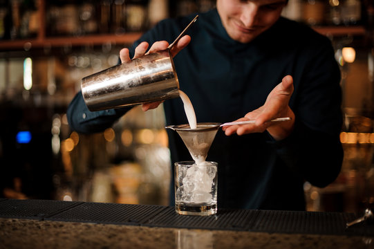 Bartender pouring an alcoholic cocktail into a cocktail glass from a steel shaker