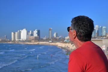 Satrszy mężczyzna z lekko siwiejącymi włosami, w czerwonej koszulce i okularach słonecznych, stoi bokiem i spogląda na panoramę zatoki Morza Śódziemnego, plażę i wyokie, nowoczesne budynki Tel Awiwu - obrazy, fototapety, plakaty