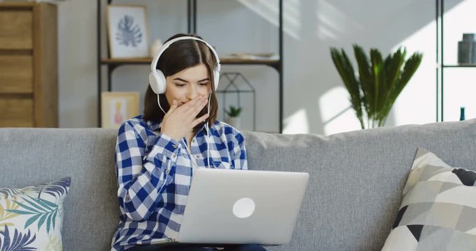 young brunette woman in the plaid blue shirt and in headphones watching something on the laptop computer screen and making scarry face. Indoor
