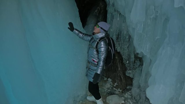 Travel woman on ice cave of Lake Baikal. Trip to winter island. Girl backpacker is walking of ice grot. Traveler looks at beautiful ice grotto. Hiker wears sports glasses, silver jacket, backpack, led