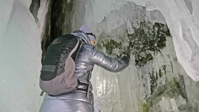 Travel woman on ice cave of Lake Baikal. Trip to winter island. Girl backpacker is walking of ice grot. Traveler looks at beautiful ice grotto. Hiker wears sports glasses, silver jacket, backpack, led