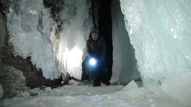 Travel man on ice cave of Lake Baikal. Trip to winter island. Backpacker is walking of ice grot. Traveler looks at beautiful ice grotto. Hiker wears sports glasses, silver jacket, backpack, led lamp