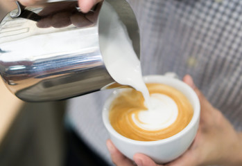 Woman barista pouring stream milk for making latte art coffee with heart shape in white cup, selective and soft focus