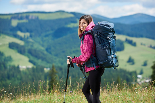 Portrait of beautiful sporty woman hiker with blue backpack and trekking sticks, hiking in the mountains, smiling to the camera, enjoying summer day in the mountains. Concept of active lifestyle