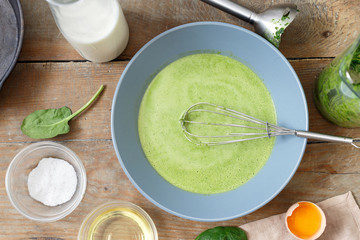 Bowl of dough with ingredients for cooking healthy spinach pancakes on wooden table top view. Gluten free spinach pancakes recipe