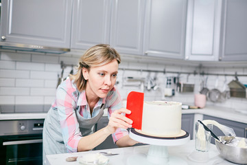 Young attractive Caucasian woman in apron carefully smoothing delicious frosted cake in kitchen